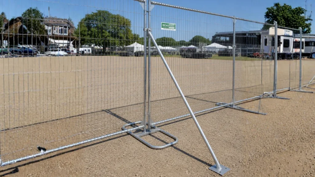 Metal temporary fence panels on a dirt field, with tents, cars, and a trailer visible in the background under a clear blue sky.
