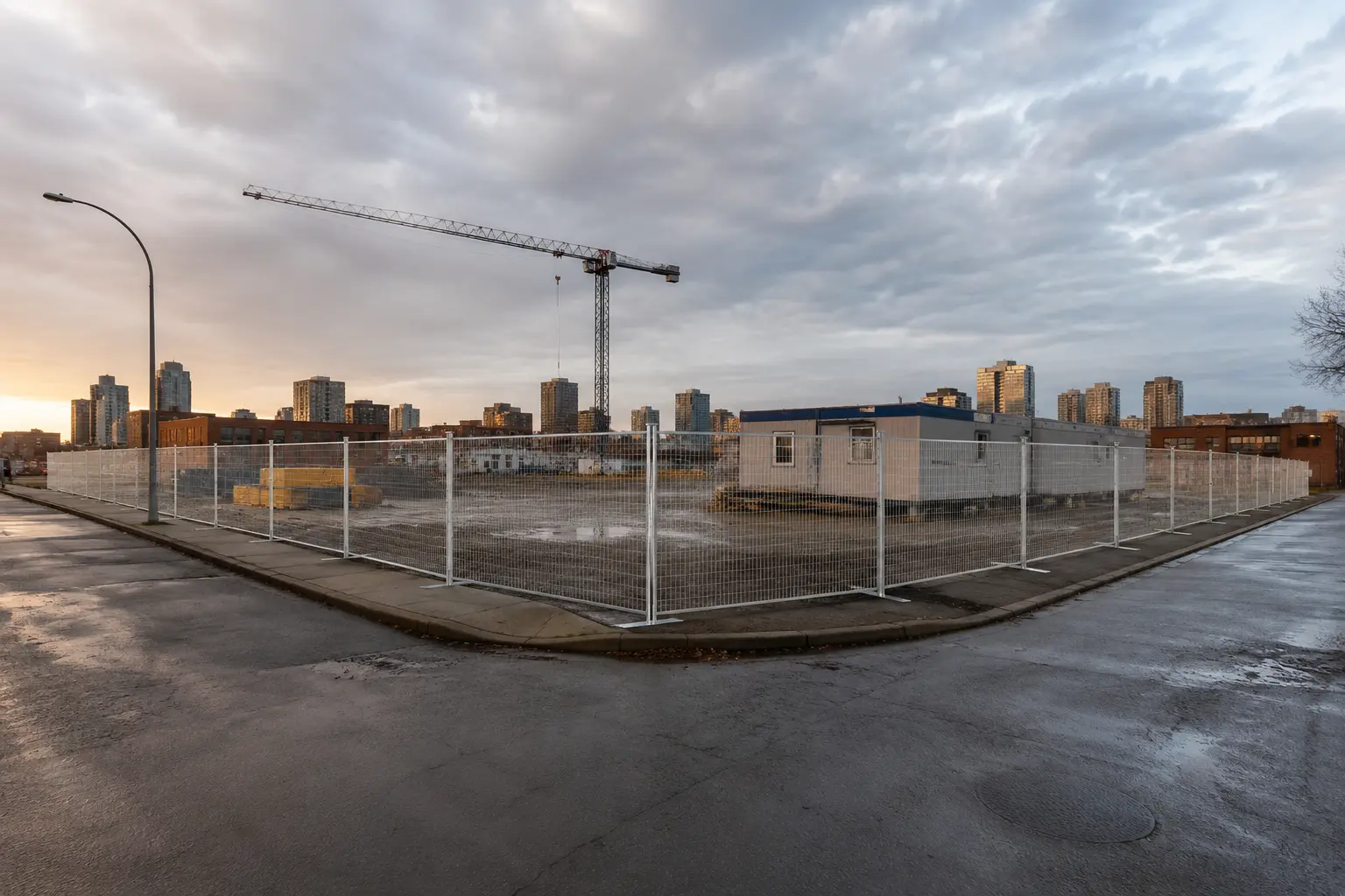 Construction site inside a white chain-link fence on a wet street at sunset, with a tall crane and city buildings in the background