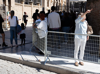 People lined up behind metal barricades on a cobblestone street, waiting near a building; one person in white shields eyes with a raised arm.
