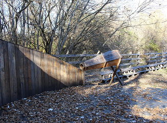 Broken wooden bench leaning against a weathered wooden fence along a leaf-littered path in a wooded area.